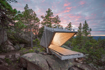 A hut or shack built with minimalist architecture stands on top of a forested hill of the High Coast of Sweden, with a colorful twilight sky behind it.
