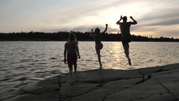 Two children and a man is jumping from a cliff into the water during a summer evening.