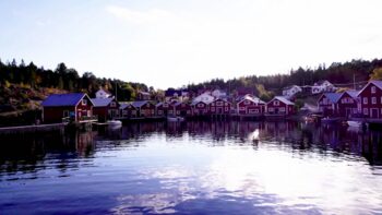 A drone flies close to water approaching the red boathouses of Bönhamn in the High Coast.