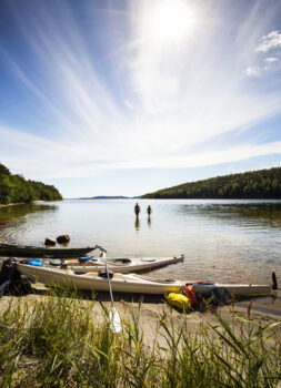 Three kayaks lay on a beach next to the water on a sunny summer day. Two people are walking in the water to take a swim. One person is standing by the kayak.