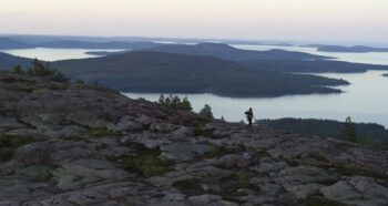 Drone footage of a woman hiking on a mountain. Behind her is a view of the High Coast archipelago.